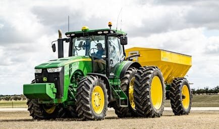 Contemporary agricultural machinery with trailer riding along grassy field in countryside against cloudy sky
